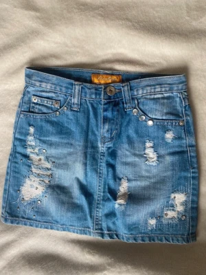 Blå jeanskjol med slitningar  - Säljer en kort blå jeanskjol med slitna detaljer och coola nitar på fickorna fram och bak. Kjolen har klassisk femficksmodell och bälteshällor. Perfekt för dig som gillar en edgy och trendig stil. W 25
