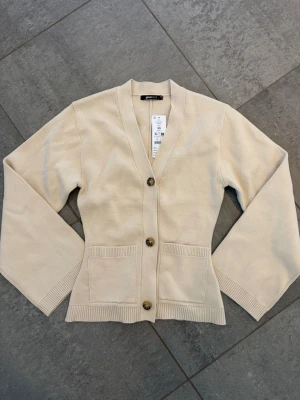 Beige kofta med fickor och v-ringning - Stilren beige kofta med v-ringning och tre knappar framtill. Koftan har två praktiska fickor och ribbade muddar vid ärmslut och nederkant. Passformen är något boxig och materialet ser mjukt och bekvämt ut, perfekt för lager på lager. Ny med prislapp, nypris 459kr