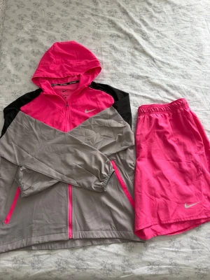 Nike Miller träningsset - Säljer ett sportigt träningsset från Nike med en tunnjacka och shorts. Jackan har dragkedja, huva och fickor, i färgerna neonrosa, grått och svart. Shortsen är neonrosa med elastisk midja och Nike-logga. Perfekt för träning eller löpning. Jackan och shortsens storlek är M