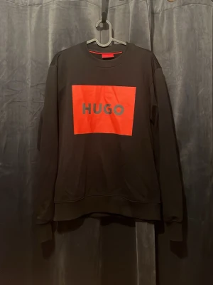Svart sweatshirt från HUGO - Svart sweatshirt från HUGO med en stor röd rektangel och HUGO-loggan tryckt på bröstet. Tröjan har rund halsringning, långa ärmar och ribbade muddar vid ärmslut och nederkant. Perfekt för en avslappnad och trendig look. Men passar också vissa S.