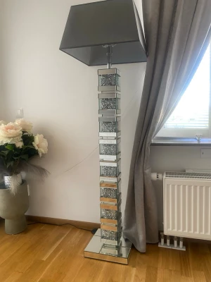 Spektakulär golvlampa med spegelglas - Unik golvlampa med fyrkantig fot och pelare i spegelglas och dekorativa kristalldetaljer. Lampan har en stor, fyrkantig grå lampskärm som ger ett modernt och lyxigt intryck. Perfekt som statement piece i vardagsrummet pris 3000 kr styck 