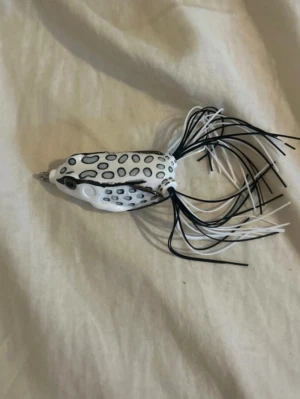 Vitt och svart fiskedrag groda - Säljer ett fiskedrag format som en groda i vitt med svarta prickar och detaljer. Draget har långa svarta och vita gummiben baktill som ger livlig rörelse i vattnet. Perfekt för dig som gillar fiske och vill testa något annorlunda i draglådan.