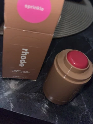 Rhode Pocket Blush Sprinkle - Krämrouge i stickformat från Rhode i färgen Sprinkle, en fräsch rosa nyans. Kommer i en smidig brun förpackning med rundad topp som gör det enkelt att applicera direkt på kinderna. Perfekt för att ge en naturlig glow och enkel att ta med i väskan.