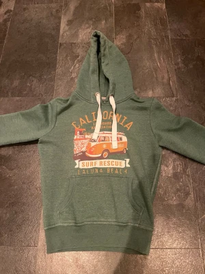 Grön hoodie med California-tryck - Snygg grön hoodie med huva och vita dragsnören. Framtill finns ett färgglatt tryck med texten 'California Surf Rescue Laguna Beach' och en retro surfvan i orange och vitt. Hoodien har känguruficka och ribbade muddar. Perfekt för chill dagar eller häng med vänner.