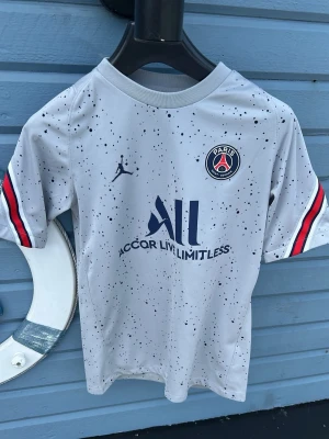 PSG vit matchtröja med stänk - Säljer en vit PSG matchtröja från Jordan med svart stänkmönster och klubbmärke på bröstet. Tröjan har korta ärmar med röd, vit och blå rand på ärmsluten samt tryck framtill. Perfekt för dig som älskar fotboll och vill sticka ut på planen.