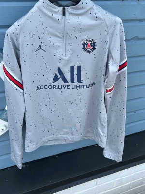 PSG x Jordan grå träningströja - Snygg grå träningströja från PSG x Jordan med svartprickigt mönster, halv dragkedja och klubbmärke på bröstet. Röd, vit och marinblå rand på ärmarna ger extra stil. Perfekt för fotbollsträning eller chill. Tillverkad i funktionsmaterial.