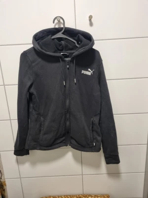 Svart hoodie från Puma med dragkedja - Säljer en svart hoodie från Puma med dragkedja framtill och två fickor. Hoodien har justerbar huva med snören och Puma-logga på bröstet. Tillverkad i mjukt material som passar perfekt för chill eller träning.