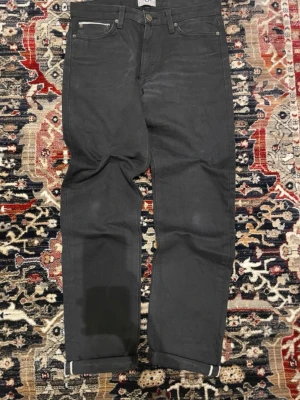 Svarta Selvedge jeans - Säljer ett par svarta jeans med rak passform och uppvikta benslut. Jeansen har klassisk femficksdesign och silverfärgade nitar. Materialet ser ut att vara kraftig denim, perfekt för en clean och stilren look.
