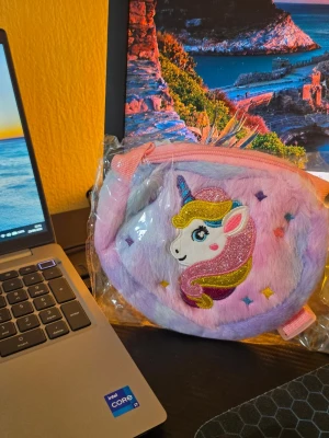 2p Rund fluffig väska med enhörning - Söt rund väska i mjukt, fluffigt material med broderad enhörning på framsidan. Väskan är ljuslila och rosa med färgglada detaljer och har dragkedja upptill samt justerbar axelrem. Perfekt för dig som gillar gulliga accessoarer och magiska motiv.