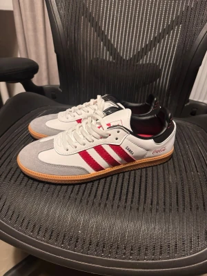 Adidas Samba x Coca-Cola sneakers - Snygga Adidas Samba sneakers i vitt och grått med röda detaljer och Coca-Cola-logga på sidan. Skorna har klassiska tre ränder, gummisula och ovandel i läder och mocka. Perfekta för dig som gillar unika samarbeten och streetstyle.