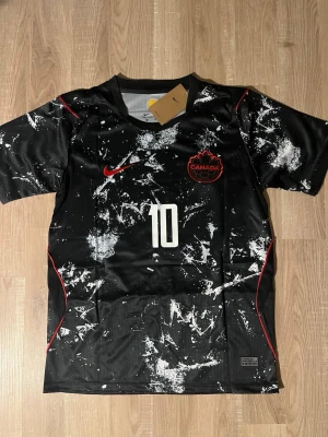 Svart Kanada fotbollströja David #10 - Snygg svart fotbollströja med Kanadas landslagsmärke och nummer 10 på bröstet. Tröjan har vitt splattermönster, röda detaljer och J. David tryckt på ryggen. Tillverkad i lätt och ventilerande material, perfekt för träning eller match. HELT NY!