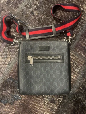Gucci axelväska med GG-mönster - Snygg svart axelväska från Gucci med klassiskt GG-mönster och en framficka med dragkedja. Väskan har en bred axelrem i rött och mörkblått, samt detaljer i silverfärgad metall. Perfekt för dig som vill ha en stilren och trendig accessoar.