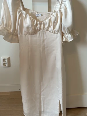 Ljusvit klänning med puffärm och slits - Elegant ljusvit klänning i satin med puffiga kortärmar och en liten slits framtill. Klänningen har en omlottdetalj vid bysten och volang vid ärmsluten, vilket ger en romantisk och trendig look.