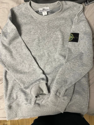 Grå sweatshirt från Stone Island - Snygg grå sweatshirt från Stone Island med klassisk rund hals och ribbade muddar. Tröjan har den ikoniska Stone Island-loggan på vänster ärm. Tillverkad i mjukt bomullsmaterial som är skönt mot huden.
