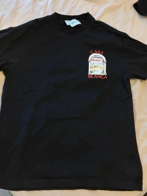 Svart Casa Blanca t-shirt med tryck - Svart t-shirt med färgglatt tryck och texten 'Casa Blanca' både fram och bak. Klassisk passform med rund hals och korta ärmar. Perfekt för dig som gillar streetwear och vill sticka ut med en unik design.