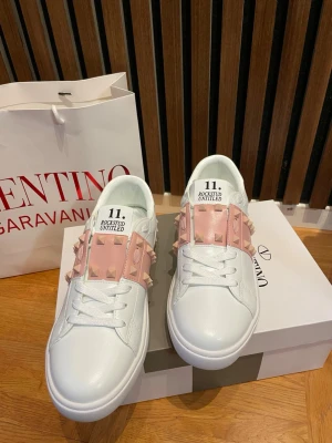 Vita sneakers med rosa nitar - Snygga vita sneakers från Valentino med breda rosa band över vristen dekorerade med matchande nitar. Skorna har en klassisk låg siluett och vita snören. Perfekta för dig som vill sticka ut med en unik detalj på dina sneakers.