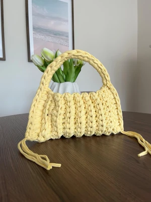 Gul virkad handväska med handtag - Superfin handväska i ljusgul färg, virkad i ett tjockt garn med ribbad struktur. Väskan har ett runt handtag och dekorativa knytband på sidorna. Perfekt accessoar för att liva upp din outfit.