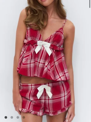 Rödvit rutigt pyjamas-set med rosetter - Pyjamas-set i två delar med rödvitt rutigt mönster. Toppen har smala axelband, v-ringning och en vit rosett framtill. Shortsen har volangkant och matchande vit rosett i midjan. Setet är gjort i ett lätt och mjukt tyg, perfekt för att chilla hemma.