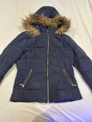 Mörkblå pufferjacka med fuskpäls - Snygg mörkblå pufferjacka med huva och fuskpäls runt kanten. Jackan har två snedställda fickor med dragkedja och markerad midja för en schysst siluett. Perfekt för kalla dagar och riktigt varm tack vare det vadderade materialet.