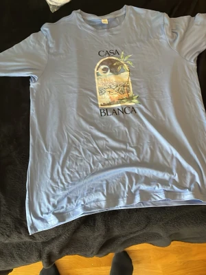 Ljusblå t-shirt med tryck Casa Blanca - Snygg ljusblå t-shirt med tryck framtill där det står 'Casa Blanca' och ett motiv med palmer och landskap. Klassisk passform och rund halsringning. Perfekt för en avslappnad stil.