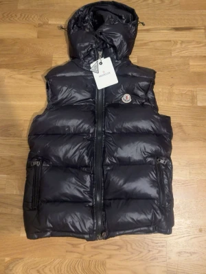 Svart dunväst med huva från Moncler - Snygg svart dunväst från Moncler med glansig finish och avtagbar huva. Västen har två dragkedjeförsedda fickor, Moncler-logga på bröstet och kraftig dragkedja framtill. Perfekt för lager-på-lager och streetwear-vibe.