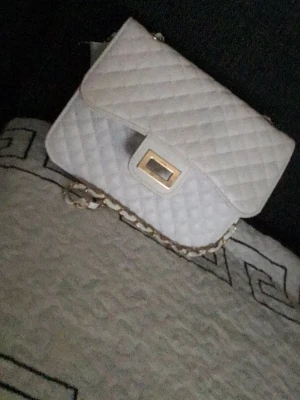 Vit quiltad axelremsväska - Snygg vit handväska med quiltat mönster och guldfärgad kedjerem. Väskan har en rektangulär form och ett elegant spänne framtill. Perfekt för dig som gillar stilrena accessoarer med en lyxig touch.
