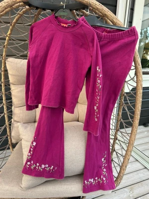 Set för flickor - Tvådelat set i lila med långärmad topp och utsvängda byxor. Båda delarna har broderade blommor i vitt och grönt längs ärmar och benslut. Toppen har volangkant vid halsen och ärmslut, vilket ger en söt och trendig look. Perfekt för dig som gillar färg och detaljer.