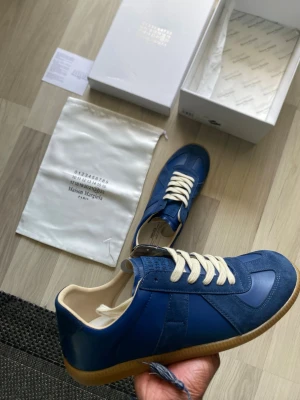 Blå Maison Margiela sneakers - Snygga blå sneakers från Maison Margiela med beige snörning och gummisula. Skorna har en mix av mocka och skinn i ovandelen och klassisk siluett. Perfekta för dig som vill ha något unikt och stilrent i garderoben. Utan tvekan helt äkta och kvitto medgår:)