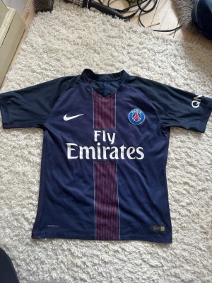 PSG matchtröja i marinblått - Snygg PSG matchtröja från Nike i marinblått med röda och vita detaljer. Tröjan har korta ärmar, klubbmärke på bröstet och Fly Emirates-tryck framtill. Tillverkad i lätt och ventilerande material, perfekt för fotbollsträning eller match.