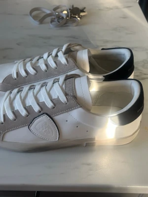 Philippe Model vita sneakers med mockadetaljer - Snygga vita sneakers från Philippe Model med beige mockadetaljer och svart hälparti. Skorna har klassisk låg profil, vita skosnören och en stilren design med märkets logotyp på sidan. Perfekta för en clean och trendig look.