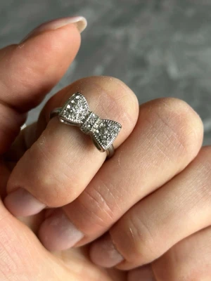 Silverfärgad ring med rosett - Söt ring i silverfärg med en glittrande rosett framtill. Rosetten är täckt av små klara stenar som ger ett elegant och lekfullt intryck. Perfekt accessoar för dig som gillar detaljer och vill sticka ut lite extra.