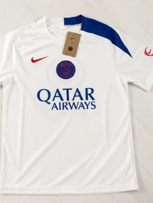 PSG matchställ Nike vit/blå/svart - Snyggt PSG matchställ från Nike med vit tröja och blå detalj på axeln, samt svart shorts med vit och blå detalj. Tröjan har PSG-logga, Qatar Airways-tryck och röd Nike-swoosh. Shortsen har elastisk midja och PSG-märke. Perfekt för fotbollsträning eller match.