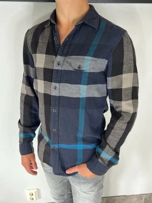 Burberry skjorta - Snygg långärmad skjorta med stora rutor i mörkblått, grått, svart och inslag av turkos. Skjortan har klassisk krage, knäppning framtill och en bröstficka med knapp. Perfekt för en avslappnad och trendig look.