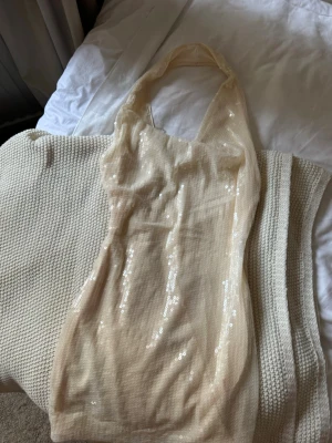 Champagnefärgad paljettklänning - Säljer en kort, champagnefärgad klänning med paljetter. Klänningen är ärmlös och har halterneck-modell. Perfekt för dig som vill sticka ut med en glittrig look. Materialet känns lätt och följsamt.