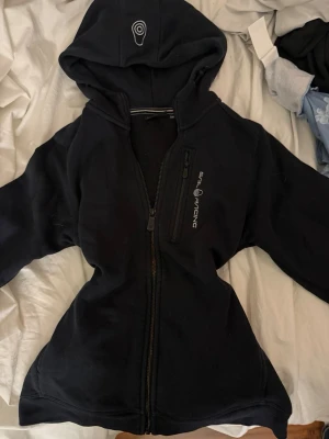 Svart hoodie från Sail Racing - Svart hoodie från Sail Racing med dragkedja framtill och en extra ficka med dragkedja på bröstet. Snygg logga på både huvan och bröstet. Perfekt för en sportig och avslappnad stil.