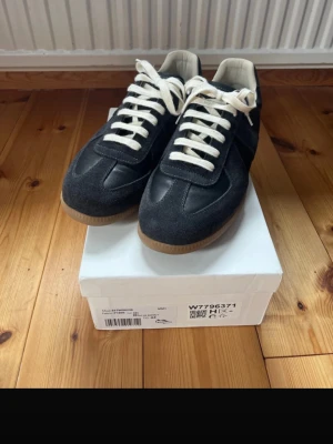 Mörkgråa Maison Margiela Gats - Snygga sneakers som endast är använda ett fåtal gånger. Svarta med grå mockadetaljer, vita skosnören och klassisk gummisula. Bekväma och stilrena – passar både till vardags och finare tillfällen. Det finns några mindre skavanker, vilket syns på bilderna. I övrigt är skorna i mycket fint skick. Säljs eftersom de inte kommer till användning.  Obs! Anledningen till att jag säljer skorna såpass billigt är pga att de är A-kopior.  Storlek: 44 (Vanlig storlek) Nypris: ca 6 000 kr