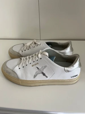 Vita sneakers från Axel Arigato -  vita sneakers från Axel Arigato med silverdetaljer och broderad fågel på sidan. Skorna har beige sula, vita skosnören och texten '1.40MM' på hälen. Tillverkade i läder .