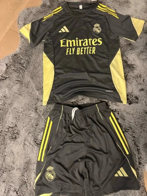 Adidas Real Madrid shortset i grön - Snygga Real Madrid shorts och tshirt  i oliv grön med gula detaljer och klubbens logga på ena benet och bröstet. Tillverkade i ett lätt och svalt material som passar perfekt för fotboll eller träning. Elastisk midja med snörning för bästa passform. Passar M bra