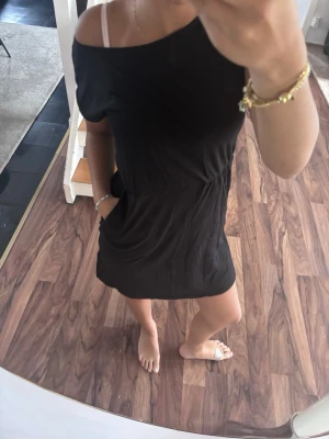 Svart kortärmad offshoulder klänning - En svart kort klänning med offshoulder-design och korta ärmar. Klänningen har en avslappnad passform och är tillverkad i ett mjukt tyg, perfekt för varma dagar. Enkel och stilren look som passar till många tillfällen.