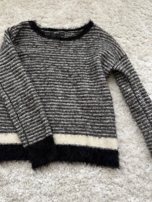 Randig stickad tröja med fluffiga detaljer - Mysig stickad tröja i svart och vitt med horisontella ränder. Tröjan har fluffiga svarta muddar vid ärmslut och nederkant samt en bred vit rand längst ner. Rund halsringning och lång ärm. Perfekt för kalla dagar och enkel att styla till jeans.
