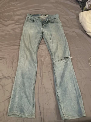 Ljusblå raka jeans från Levi's - Ljusblå jeans från Levi's med raka ben och klassisk femficksdesign. Jeansen har slitningar och hål på ena benet för en trendig, avslappnad look. Tillverkade i denim med Levi's ikoniska bakfickor och läderpatch i midjan.