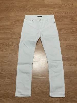 Vita Nudie jeans - Säljer ett par vita jeans från Nudie Jeans Co med klassisk femficksmodell och raka ben. Jeansen har silverfärgade nitar och knapp, samt bälteshällor. Perfekta för dig som vill ha en clean och stilren look.