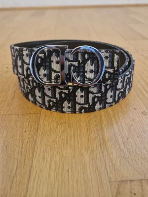 Svart och vit mönstrat bälte - Snyggt bälte med svart och vitt mönster och en stor, silverfärgad metallspänne. Bältet har ett grafiskt tryck längs hela bandet och passar perfekt för att lyfta vilken outfit som helst.