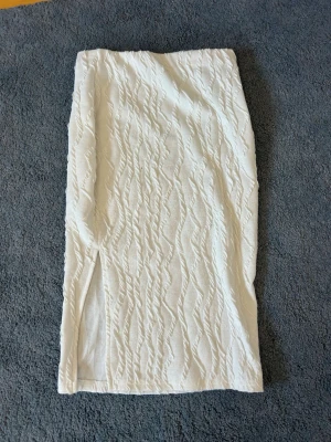 Vit långkjol med struktur och slits - Säljer en vit långkjol med vågig struktur och en snygg slits på sidan. Kjolen är gjord i polyester med lite stretch för en bekväm passform. Perfekt för dig som gillar stilrena och trendiga plagg.
