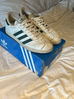 Adidas Gazelle vita och gröna sneakers - Säljer ett par klassiska Adidas Gazelle sneakers i vitt med gröna detaljer och de ikoniska tre ränderna på sidan. Skorna har låg profil, snörning och ovandel i mocka. Perfekta för dig som gillar retrostil och streetwear.