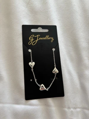 Silverfärgat halsband med hjärtan - Snyggt halsband i silverfärg från G Jewellery med tre små hjärtan som hängsmycken. Kedjan är tunn och elegant, perfekt för att ge en söt touch till din outfit. Justerbart spänne för att passa olika längder.