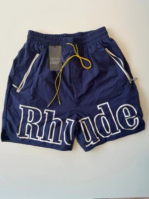 Rhude Shorts Marinblå - Rhude shorts i marinblått med stort vitt “Rhude” tryck fram, gul dragsko i kontrast och silverfärgade dragkedjor. Snygg streetwear-look, avslappnad passform.   Skick: Nyskick, prislapp kvar  Storlek: L  Skickas snabbt och säkert! 📦