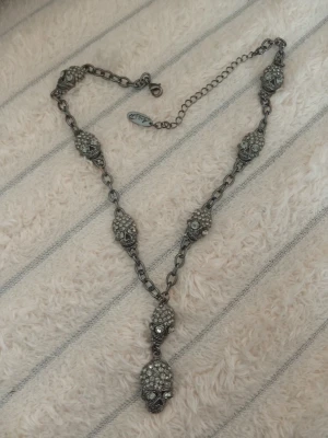 Kedjehalsband med dödskallar och stenar designat av Laila Bagge  - Unikt halsband i mörk metall med flera dödskallehängen dekorerade med små glittrande stenar. Kedjan har en cool, robust look och detaljerade dödskallar som ger en edgy vibe. Perfekt för dig som gillar statement-smycken och vill sticka ut. Halsbandet är designat av Laila Bagge 