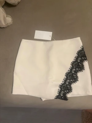 Vit skort med svart spets från Zara - Supersnygg vit skort från Zara med unik svart spetsdetalj på framsidan. Skorten har en rak passform och är tillverkad i ett slätt tyg som ger en clean look. Perfekt för dig som vill ha en trendig och stilren outfit.