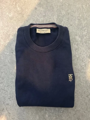 Mörkblå tröja från Burberry - Mörkblå rundhalsad tröja från Burberry med broderad logga på bröstet. Tröjan har ribbade muddar vid ärmslut och nederkant samt en klassisk passform. Halsringningen har en snygg detalj med ränder i flera färger. Perfekt för en stilren look.